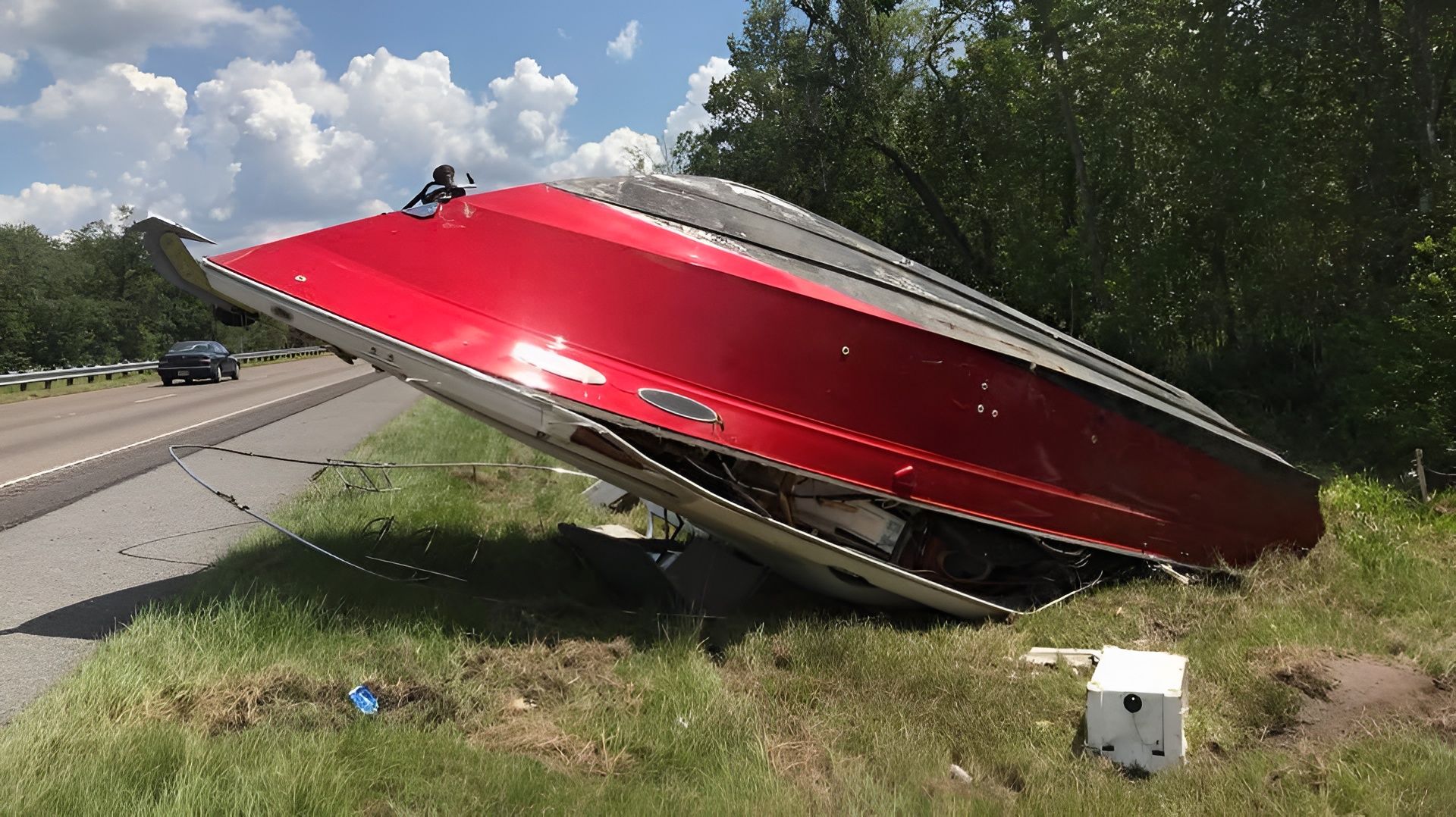 Red boat flipped on its side on the side of a road, near a guard rail and greenery.