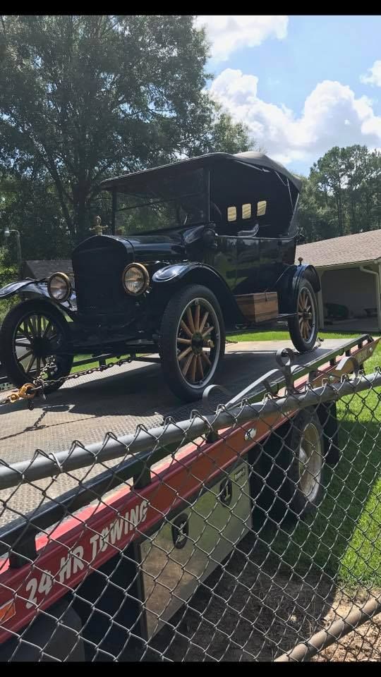 Black antique car on a tow truck, roadside. Dark wooden wheels and a canvas top.