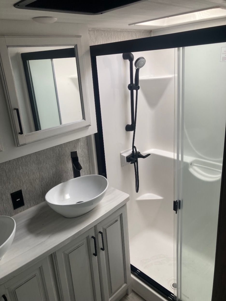 Bathroom with a white sink, shower, and vanity in an RV. Black shower fixtures and accents.