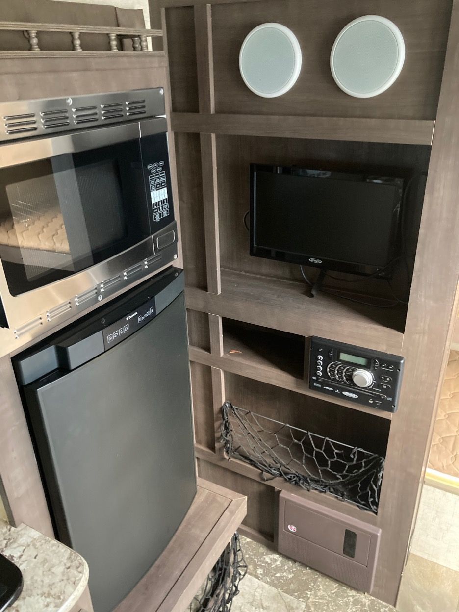 RV interior with a microwave, refrigerator, TV, radio, and storage. Light wood and gray appliances.