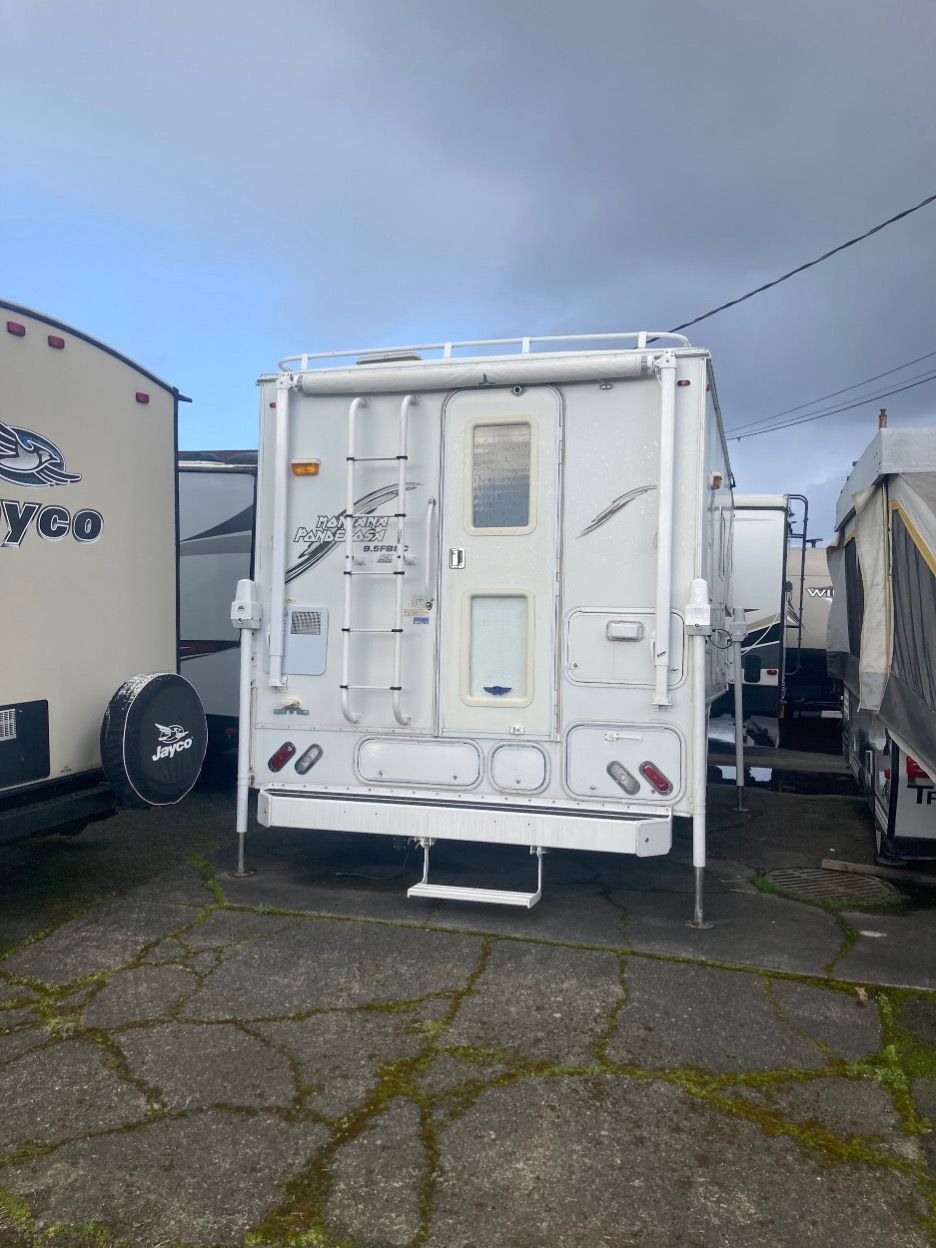 White truck camper with ladder and rooftop rack, parked among other RVs on concrete.