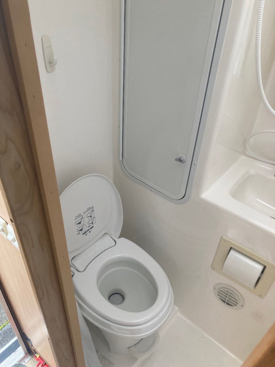 Small RV bathroom with open toilet, shower door, and sink. White walls and fixtures.