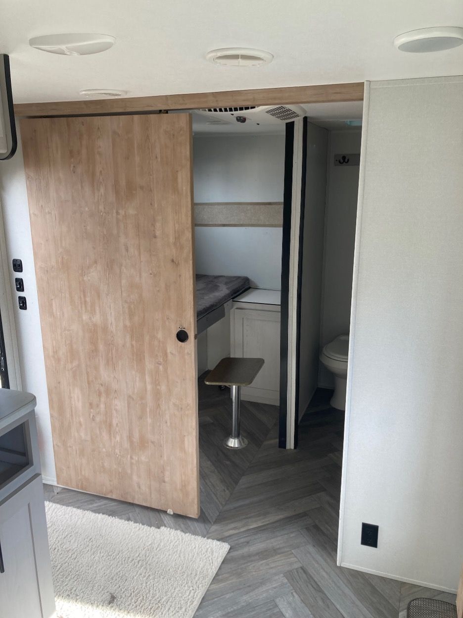 RV interior with sliding door to bedroom and bathroom; light wood, gray walls, herringbone floor.
