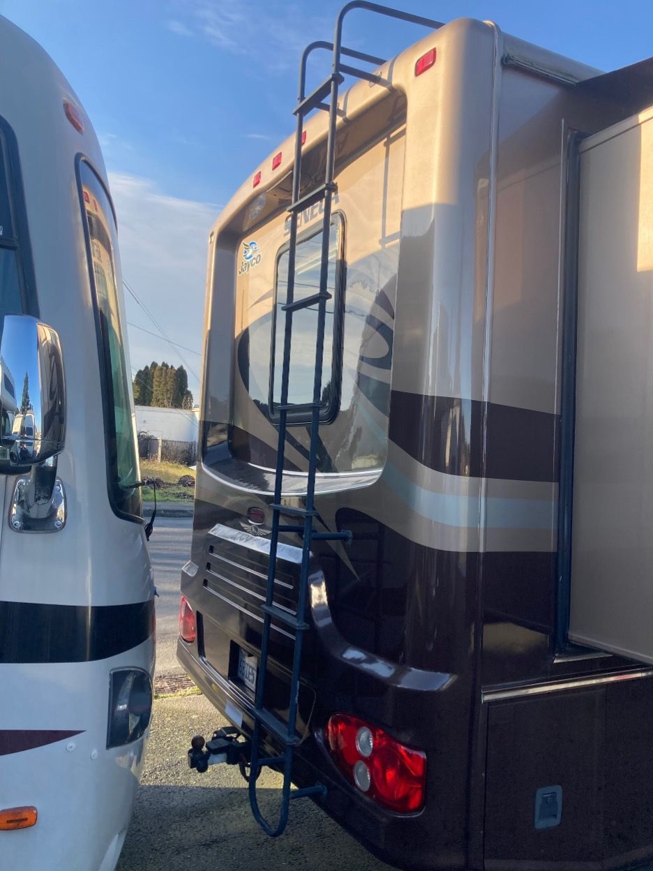 Rear view of a brown and tan RV with a black ladder, tow hitch, and tail lights.