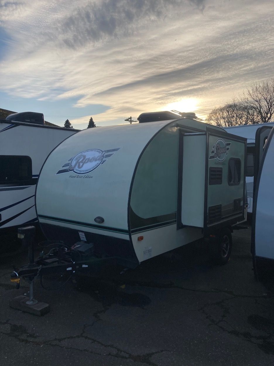 Small, light-colored travel trailer with an open door, parked outside under a cloudy sky.