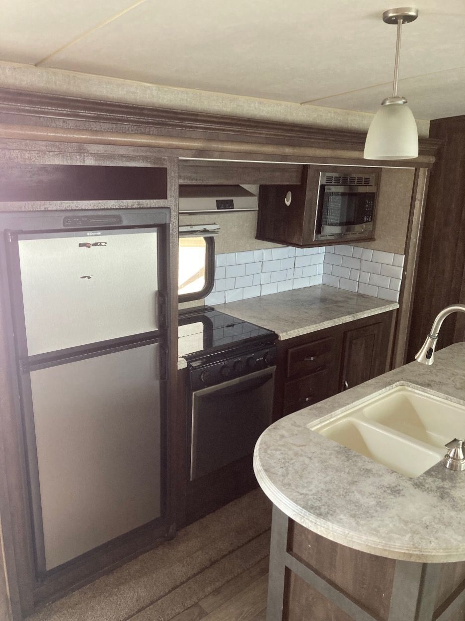 RV kitchen: refrigerator, stove, microwave, sink, and overhead light. Dark wood cabinetry and light countertops.