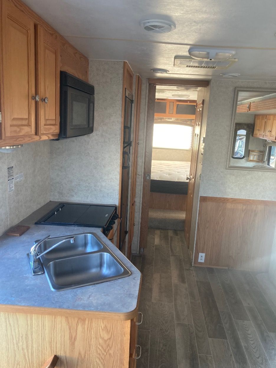 Interior of a camper trailer with kitchen and hallway leading to bedroom. Wooden cabinets and flooring.