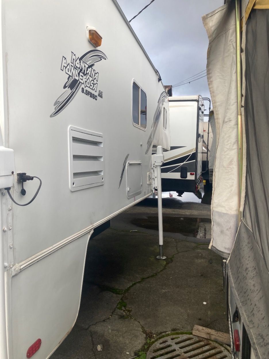 White camper side view with logo, vents, and support legs. Parked next to another vehicle.