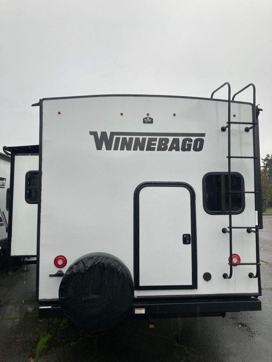 Rear view of a white Winnebago travel trailer with a ladder, spare tire, and door.