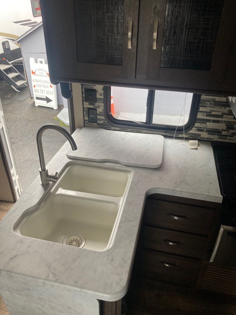RV kitchen with sink, countertop, and cabinets. Light-colored sink and counter with a window.