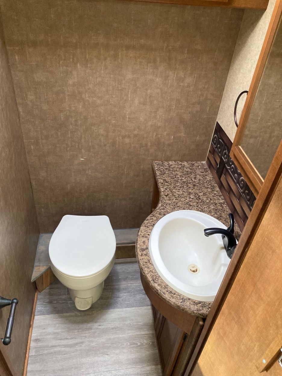 Small RV bathroom with a toilet, sink, and mirror. Brown walls, speckled countertop, and wood-tone cabinet.