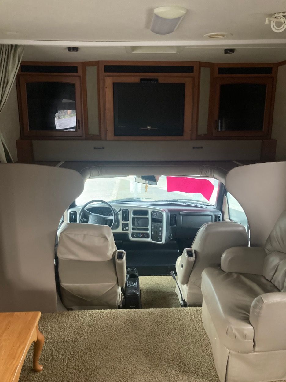 Interior of a recreational vehicle. Beige seating, dashboard, and overhead cabinets with a TV.