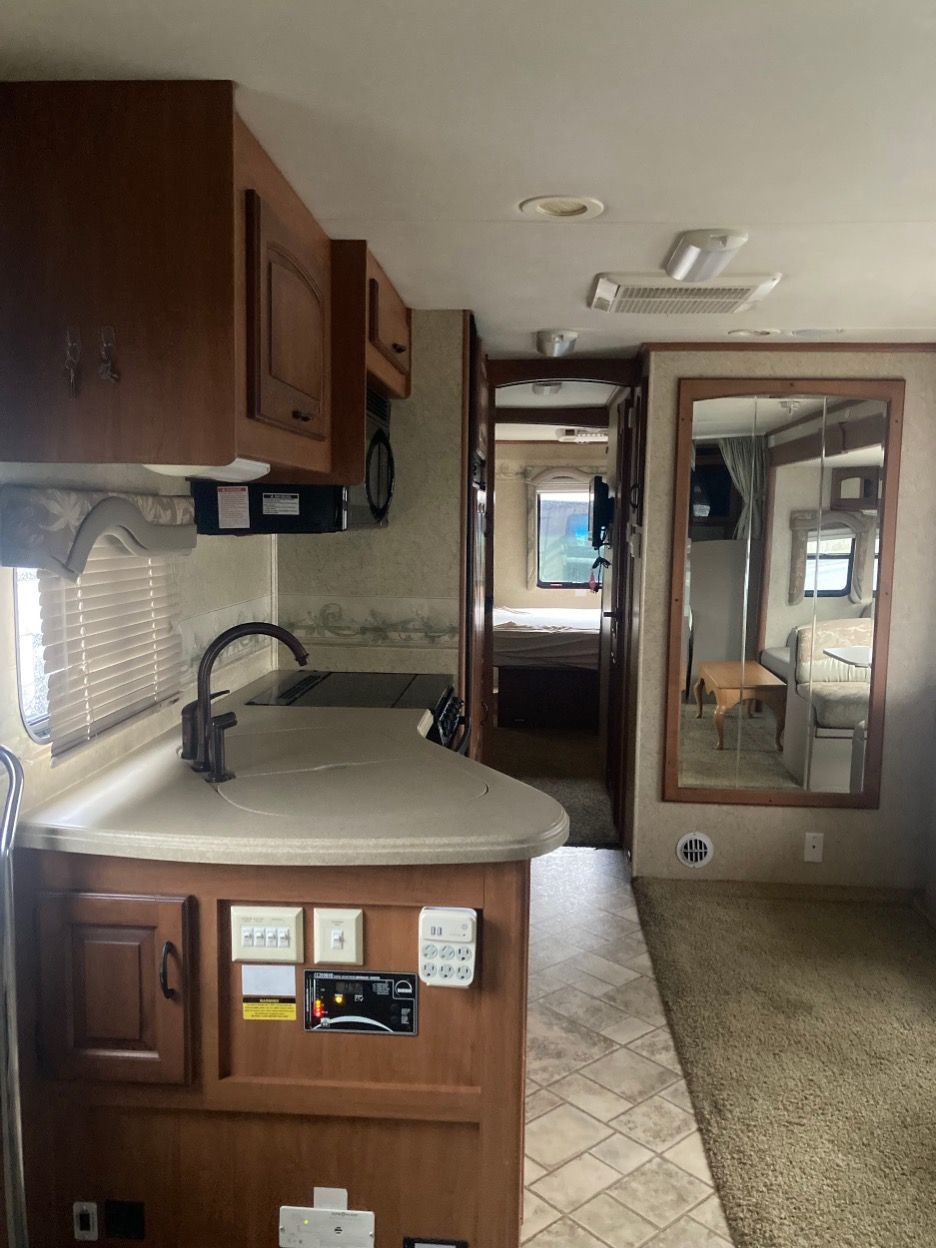 Interior view of an RV kitchen, showing a countertop, cabinets, and a doorway leading to a bedroom and living area.