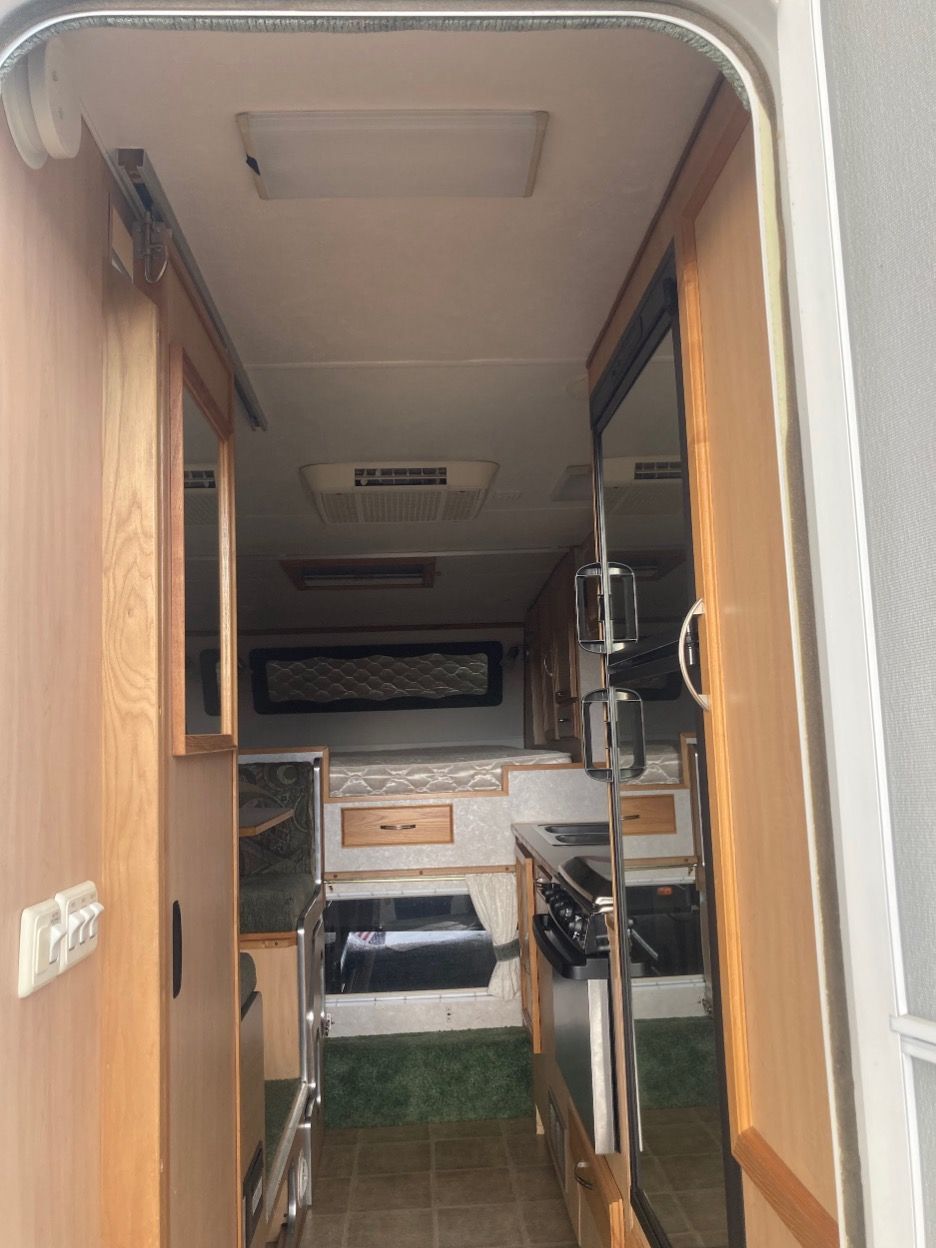 Interior view of a small RV, looking down the central aisle. Includes kitchen and bed area, wood and beige tones.