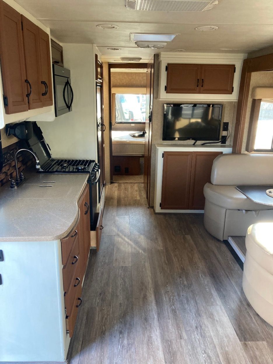 Interior of a motorhome, showing kitchen, living area with sofa and TV, and hallway leading to a bedroom.
