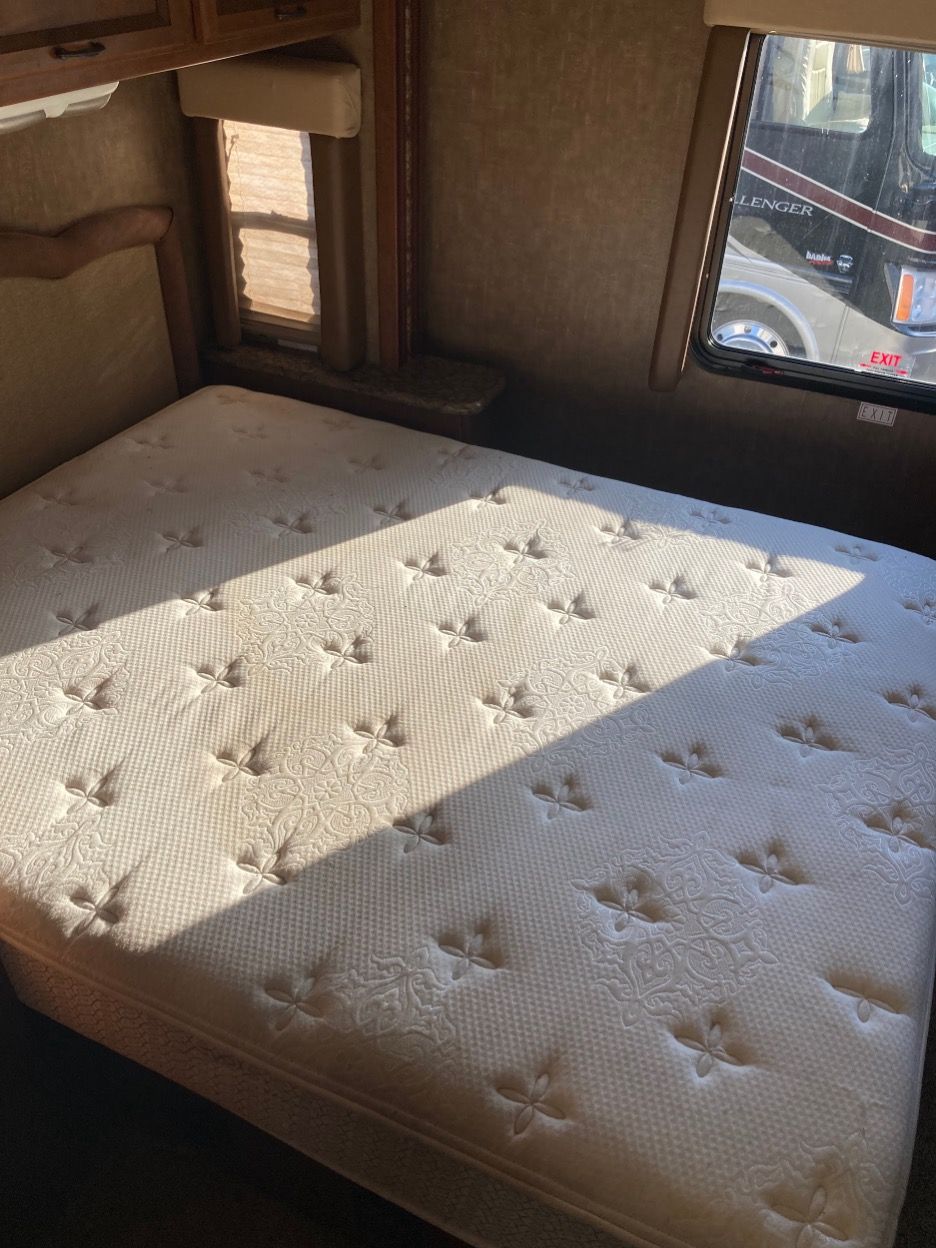 Inside an RV bedroom, a mattress with a stitched pattern rests under sunlight.