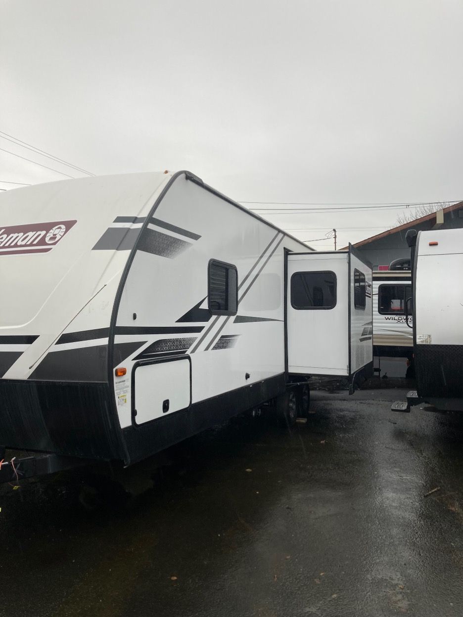 White and black Coleman RV with an open door parked on wet pavement. Cloudy sky in the background.
