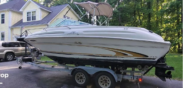 White boat on a trailer in front of a yellow house. The boat has a canvas top and gold accents.