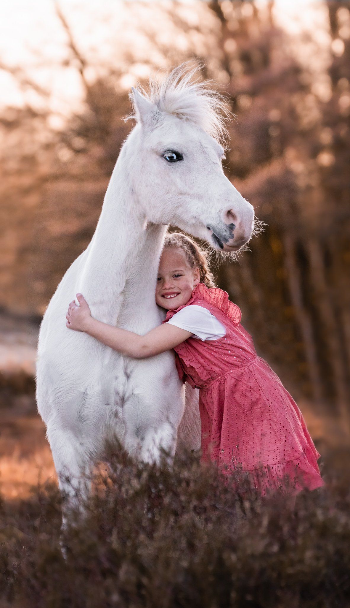 Een klein meisje in een roze jurk knuffelt een wit paard.