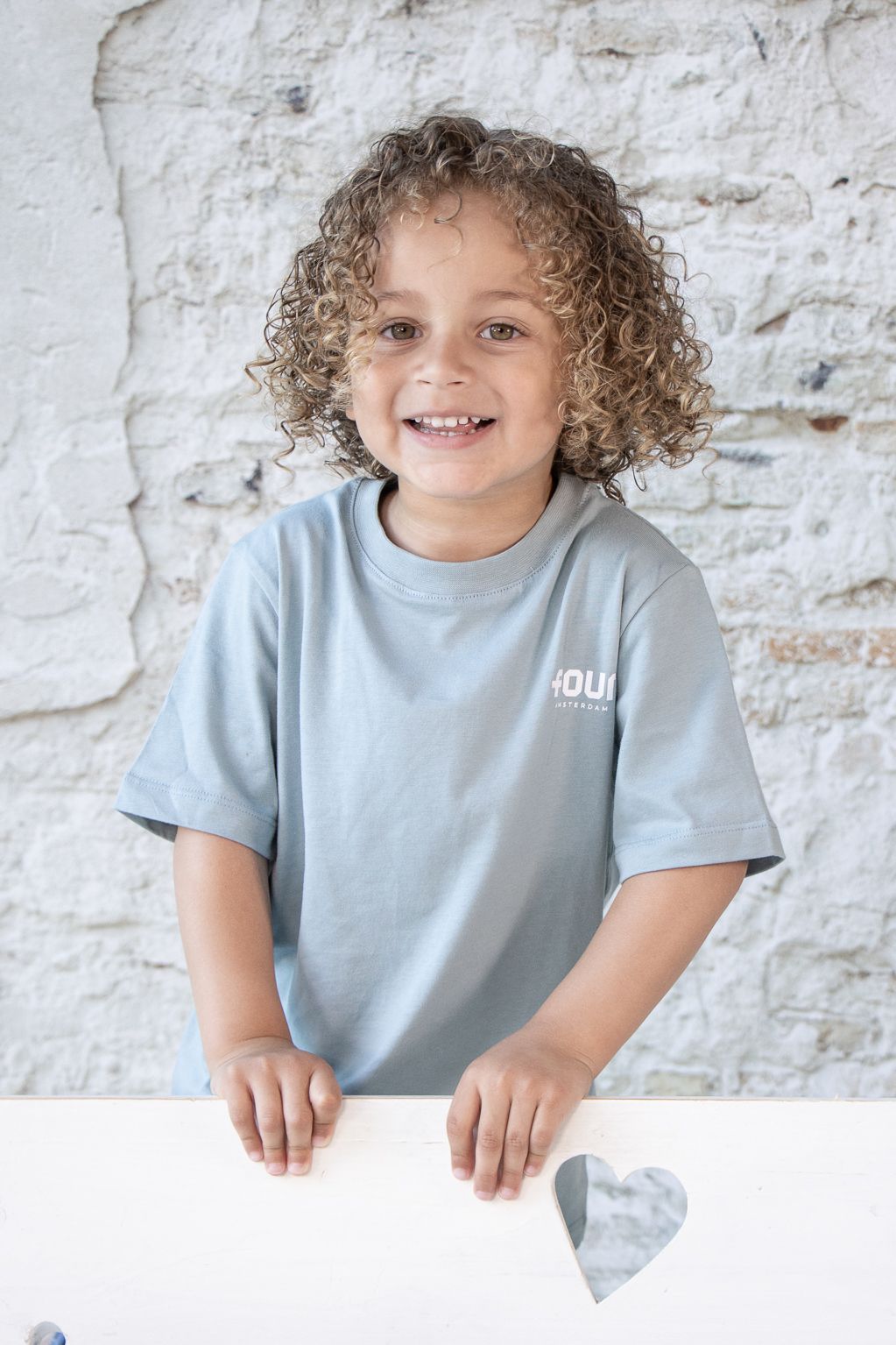 Een jonge jongen met krullend haar draagt een blauw t-shirt en glimlacht.
