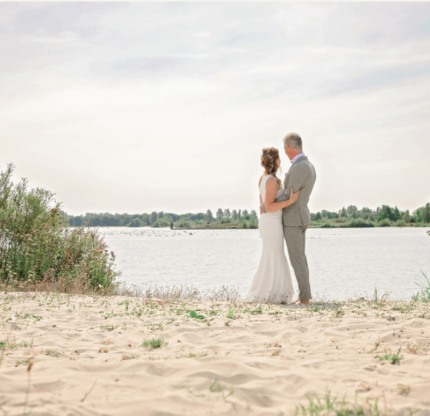 Een bruid en bruidegom staan op een zandstrand vlakbij een watermassa.