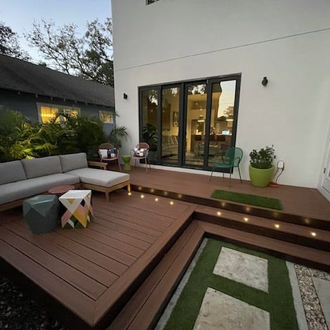A modern outdoor deck features a sectional sofa, a geometric side table, and steps leading down to a green grass path.