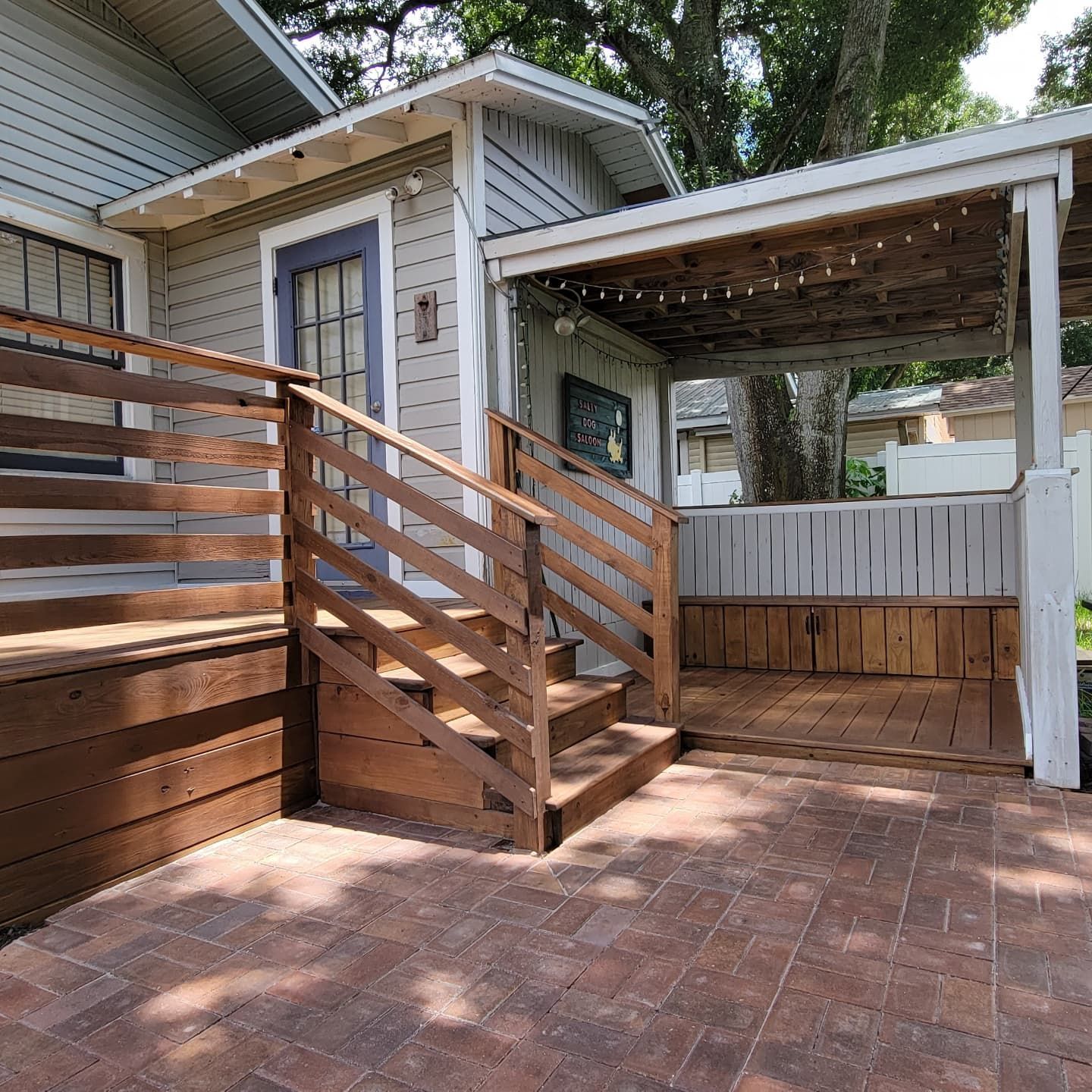 A wooden deck with a staircase leads to the side entrance of a grey house, featuring a covered patio with a wooden bench.