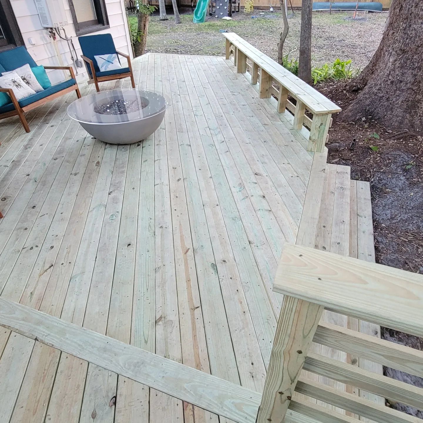 A newly built wooden deck features two lounge chairs, a fire pit, and a long built-in bench near a tree in a backyard.