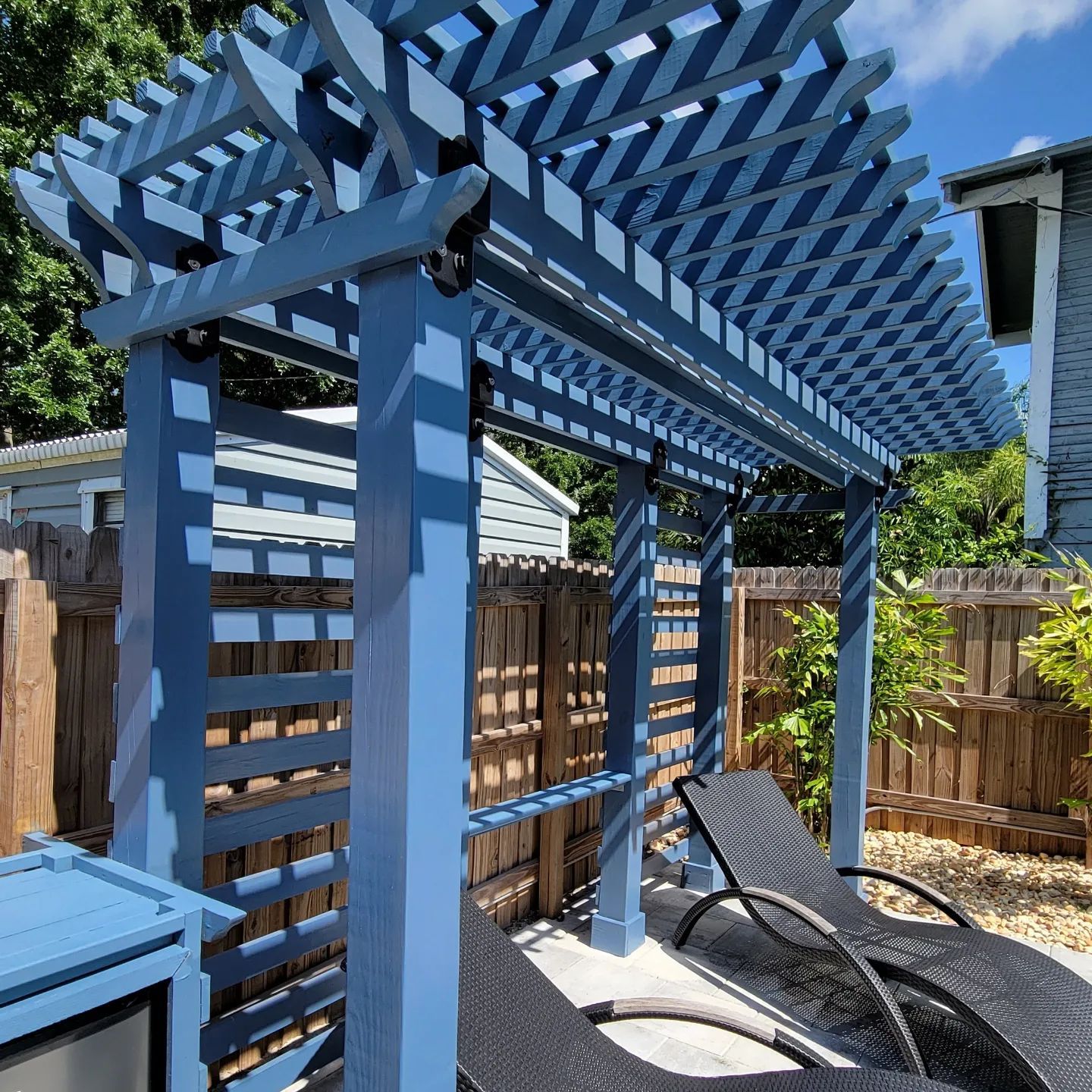 A blue wooden pergola stands over two outdoor lounge chairs in a backyard setting with a wooden fence.