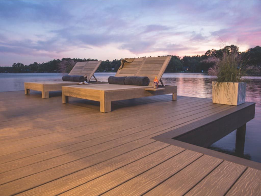 Two wooden lounge chairs sit on a modern deck over a lake at sunset, next to a planter with tall grass.
