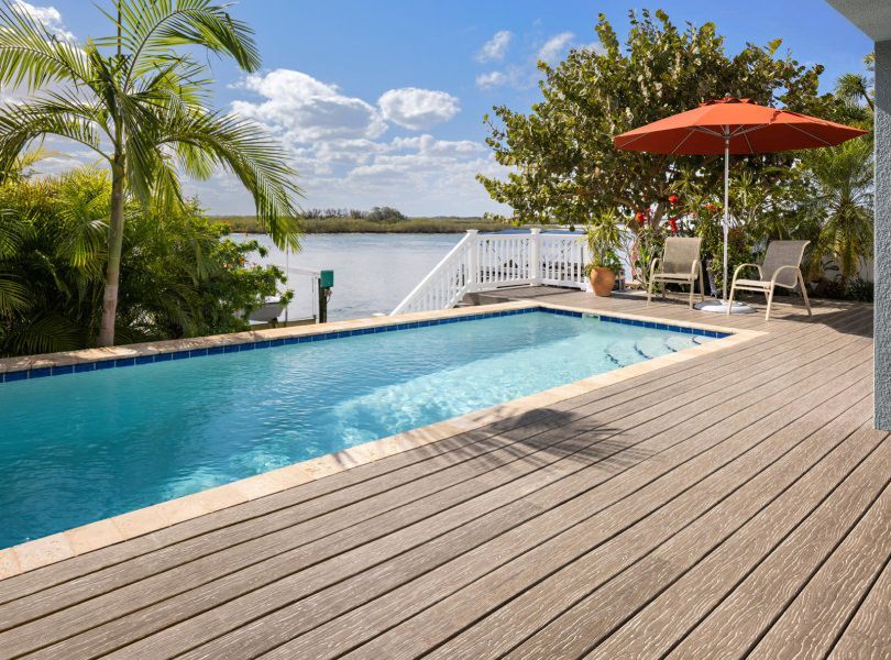 A swimming pool and wooden deck overlooking a calm body of water with palm trees and an orange patio umbrella.