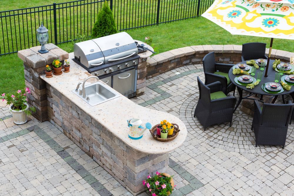 Outdoor kitchen with grill, sink, and seating area under an umbrella on a brick patio.