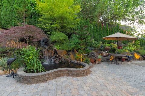 Backyard patio with a pond, waterfall, umbrella, and table surrounded by lush greenery and stone pavers.