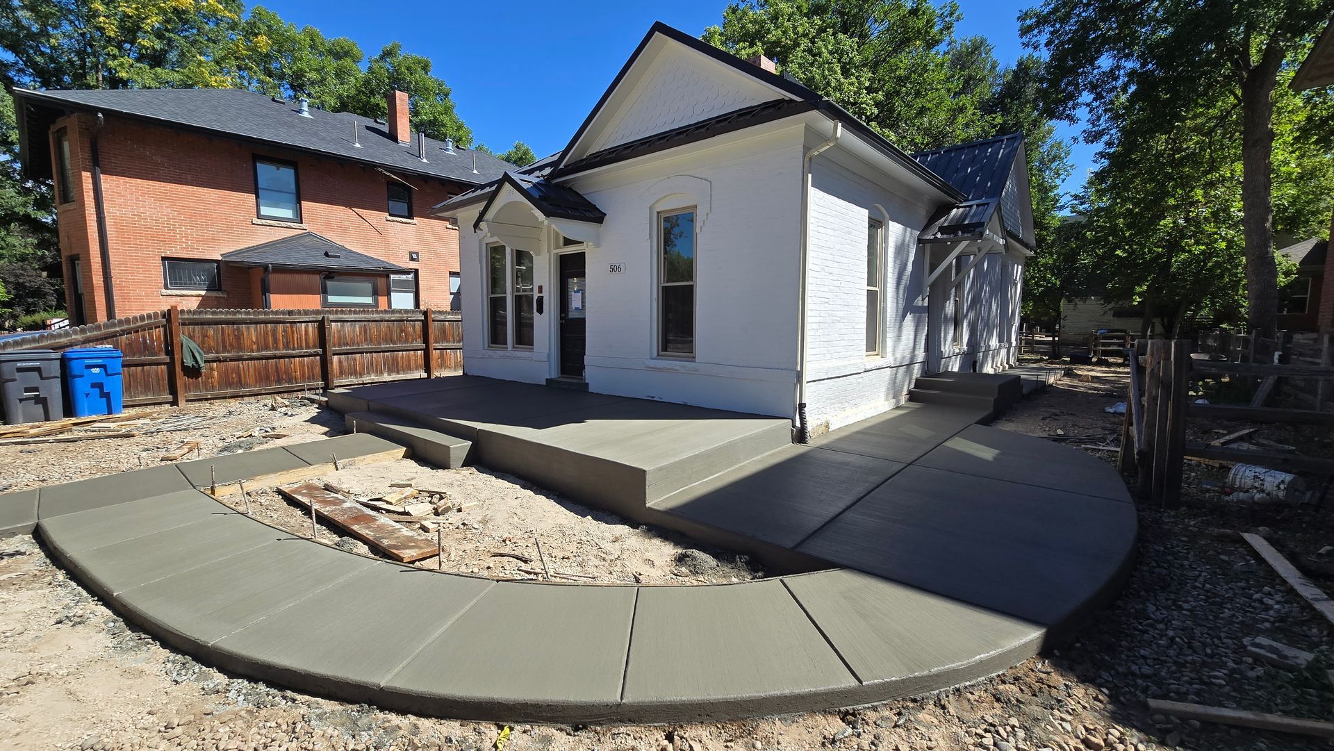 Backyard patio with curved concrete walkway, small white house, and brick building under trees