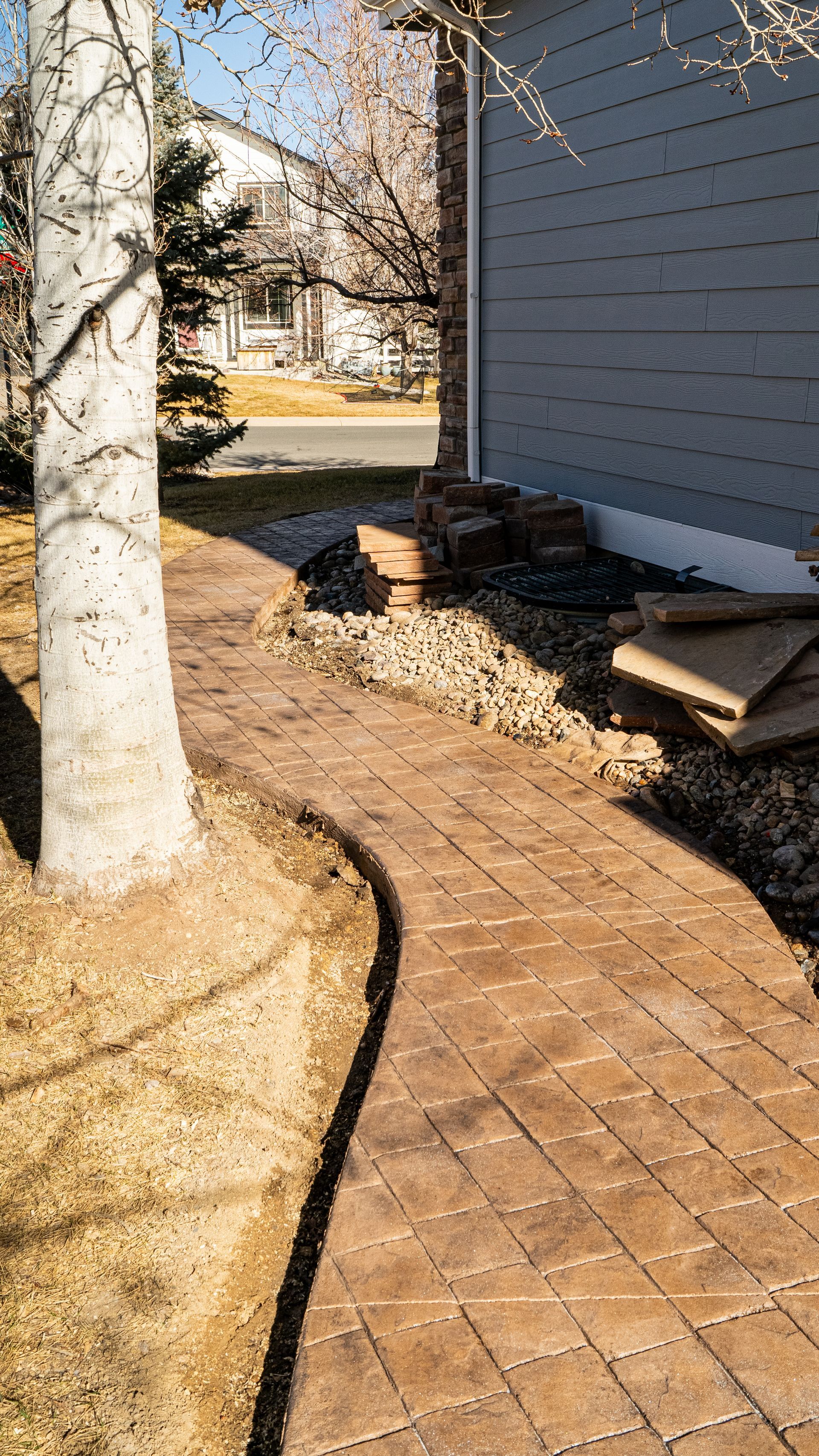 Brick walkway beside a house, with a tree trunk in the foreground and yard debris along the wall.