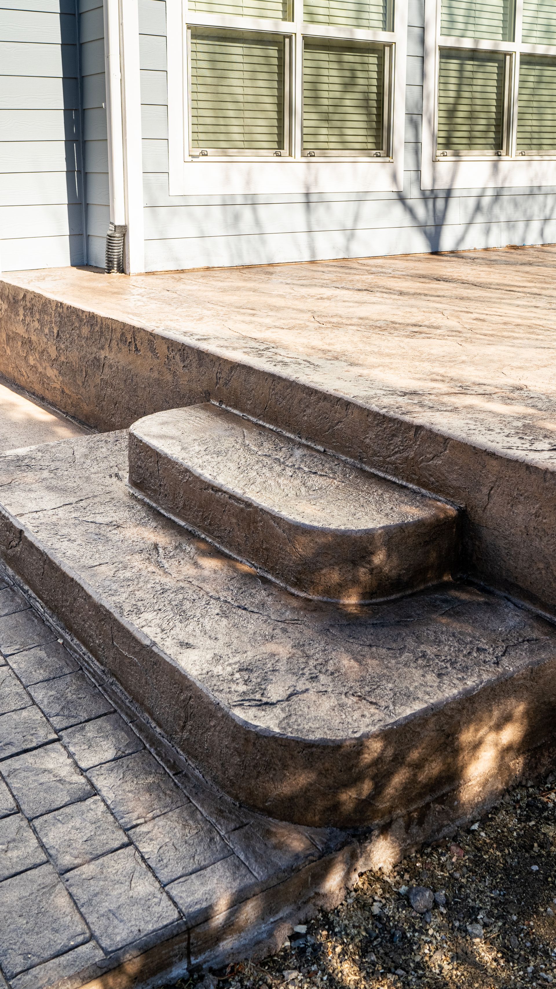 Exterior concrete steps leading to a raised patio beside a house with windows and paving stones