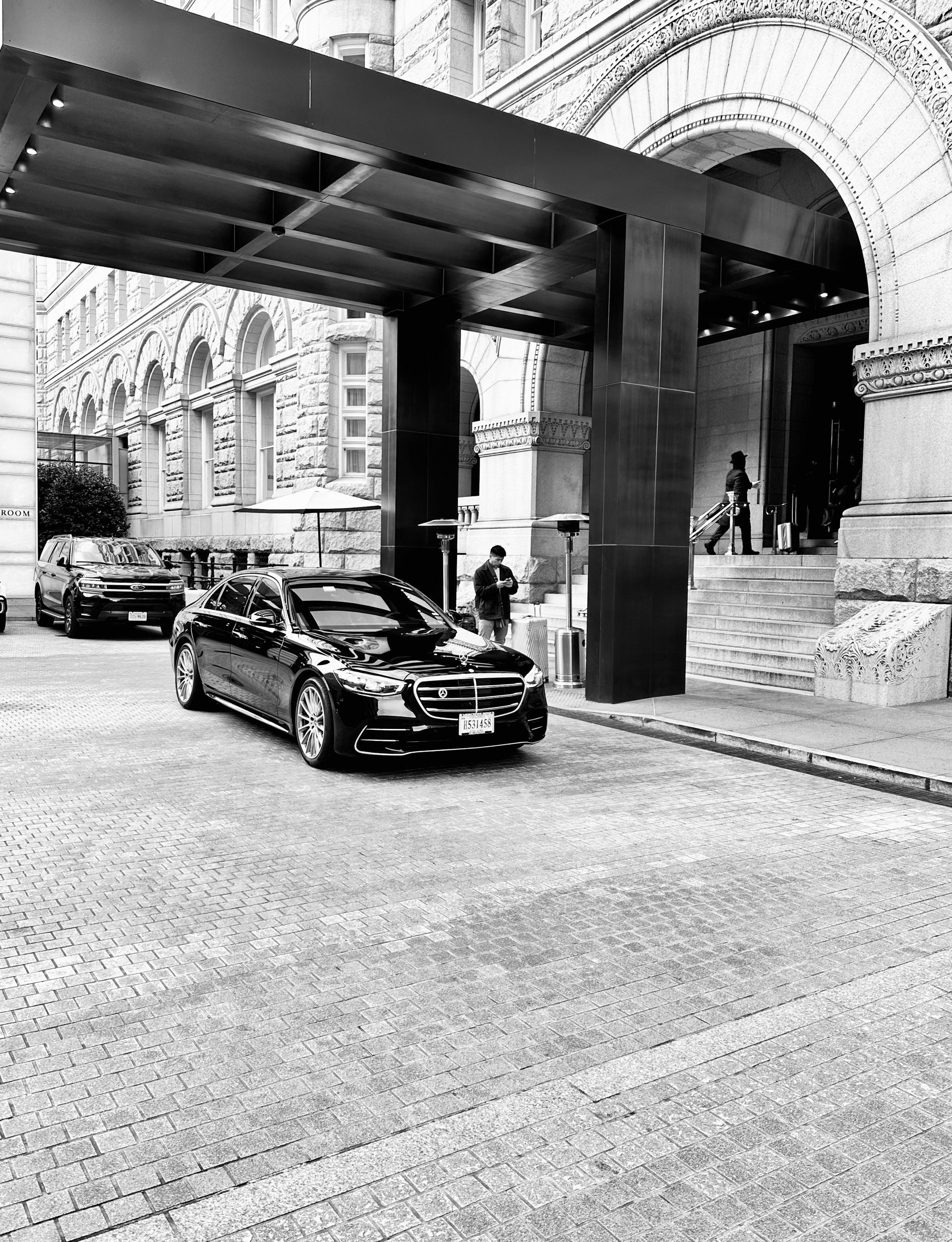 A black and white photo of a car parked under a canopy in front of a building.