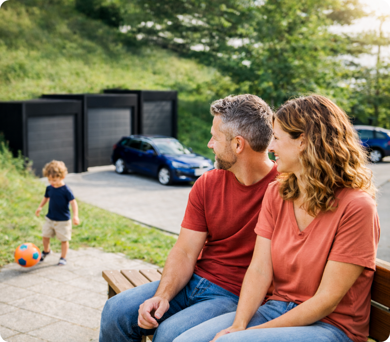 A couple sits on a park bench watching a child play with a soccer ball in front of modern garages and a parked car.