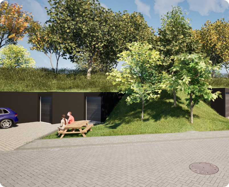 A pair sits at a wooden picnic table next to a dark wall partially buried in a grassy, tree-lined berm by a paved lot.