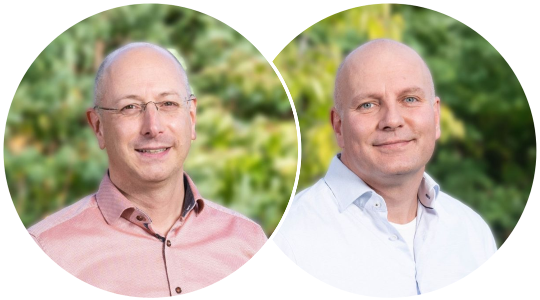 Two men in button-down shirts, one pink and one white, smiling in front of a blurry outdoor green background.