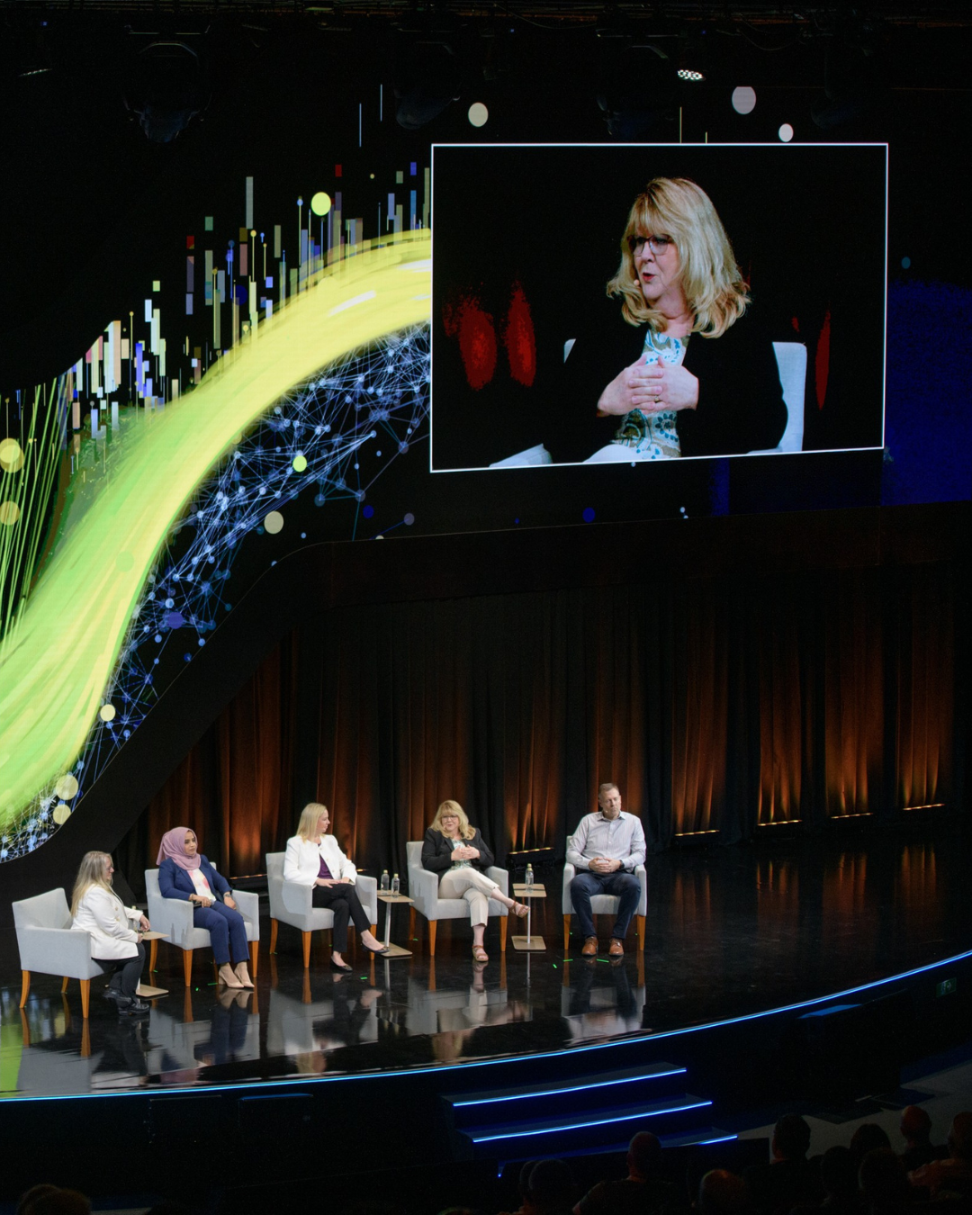 A group of people sitting in chairs on a stage with a large screen behind them