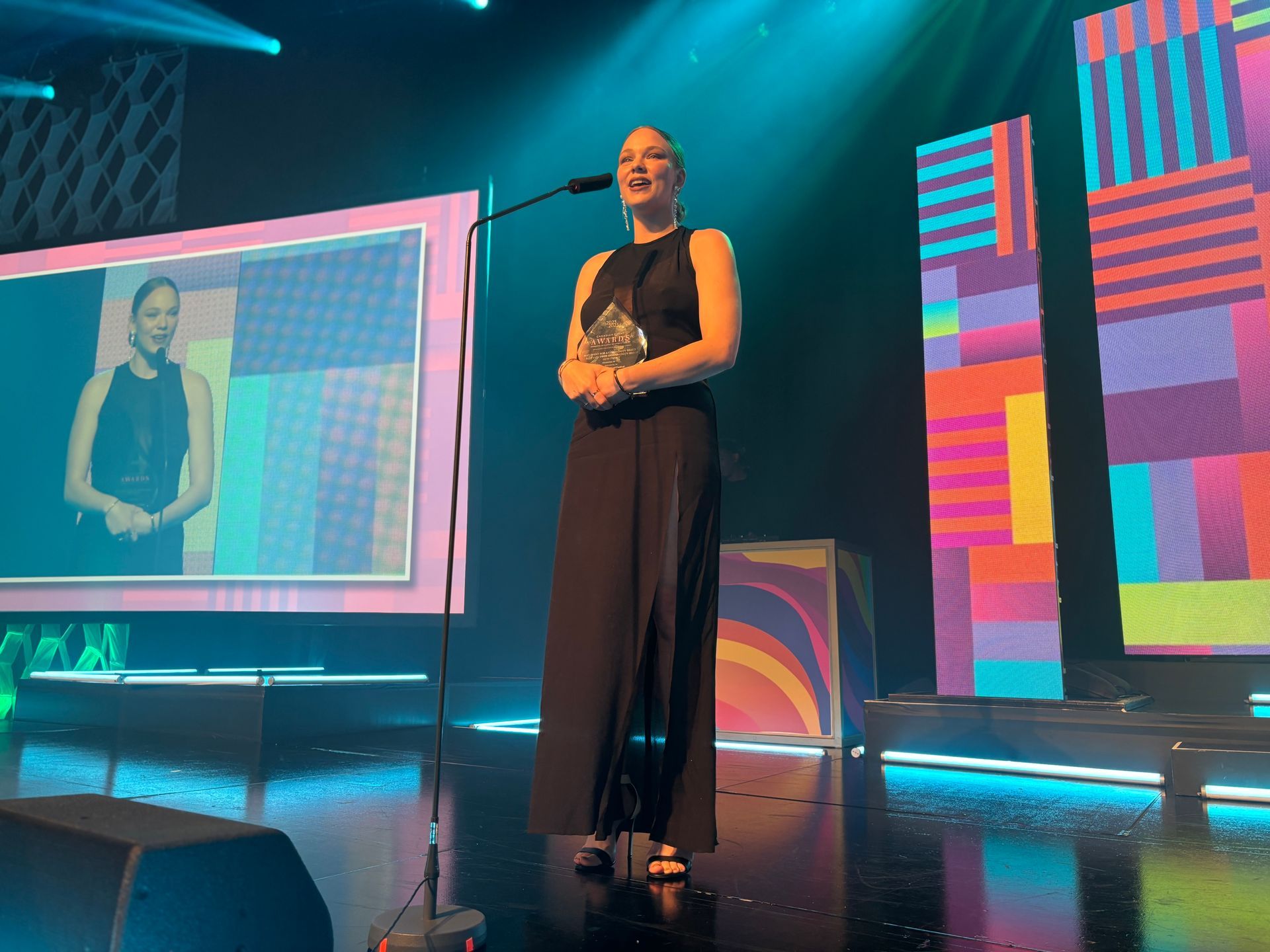 Woman on stage in black dress, holding award. Stage set with screens and colorful geometric designs.