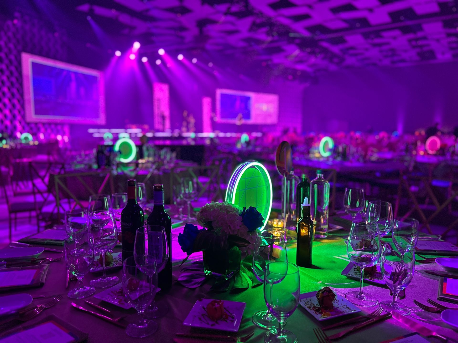 Tables set for an event, lit with purple and green lights, stage in the background.
