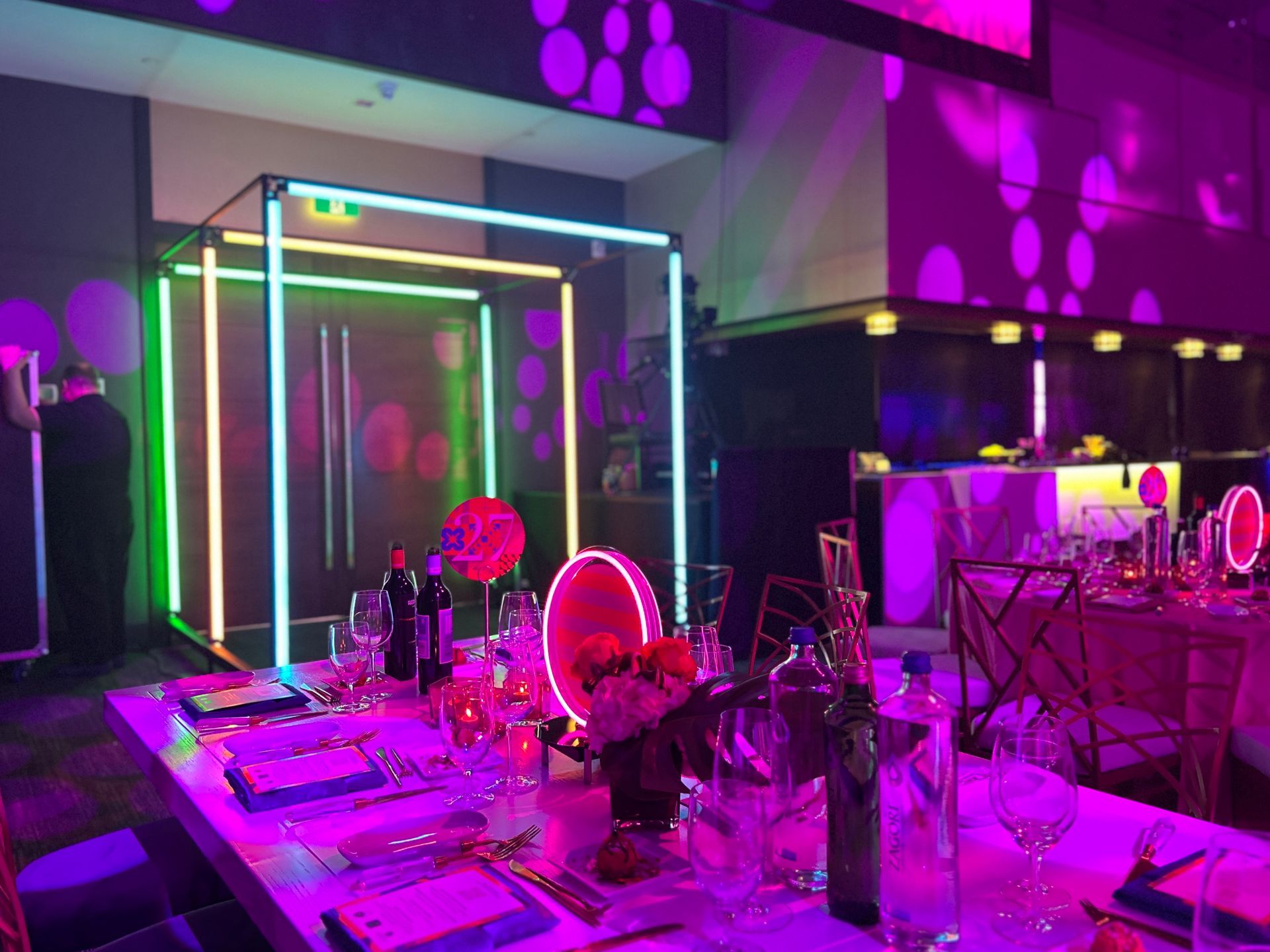 A dining table set with glasses and bottles, with neon light structures and purple/green lighting in a room.