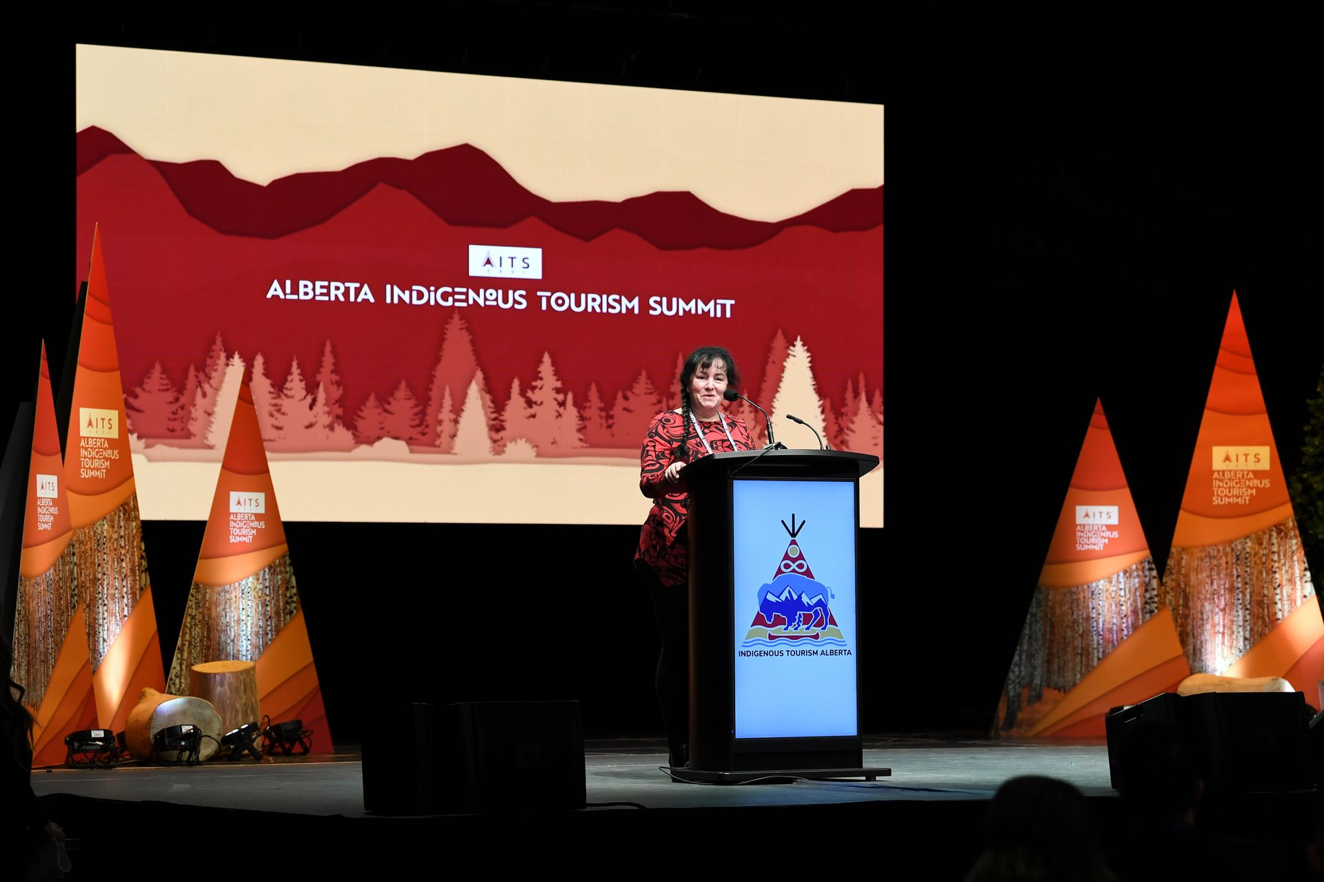 A woman stands at a podium in front of a large screen that says alberta indigenous tourism summit
