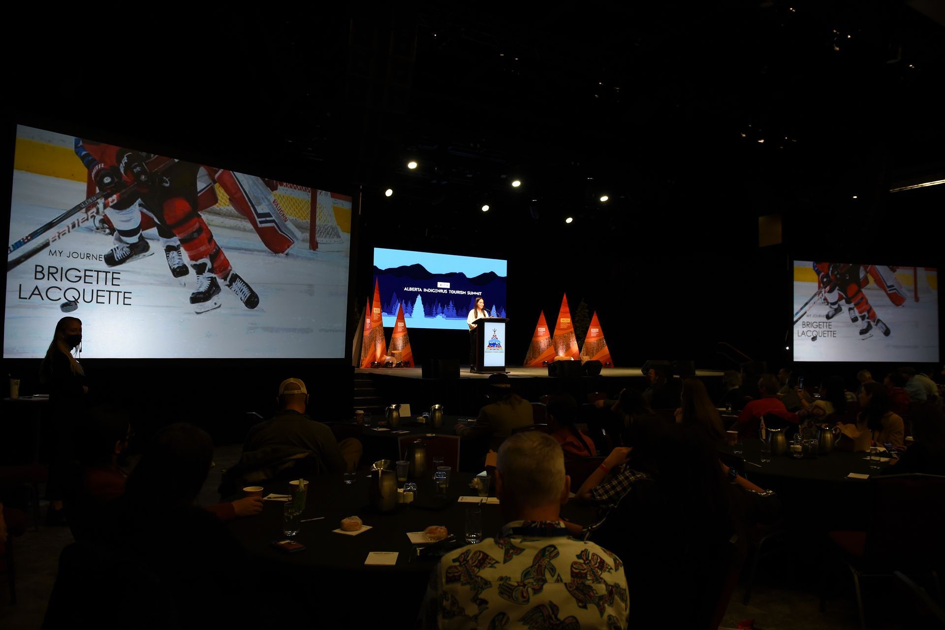 A group of people are sitting at tables in a dark room watching a hockey game on a large screen.