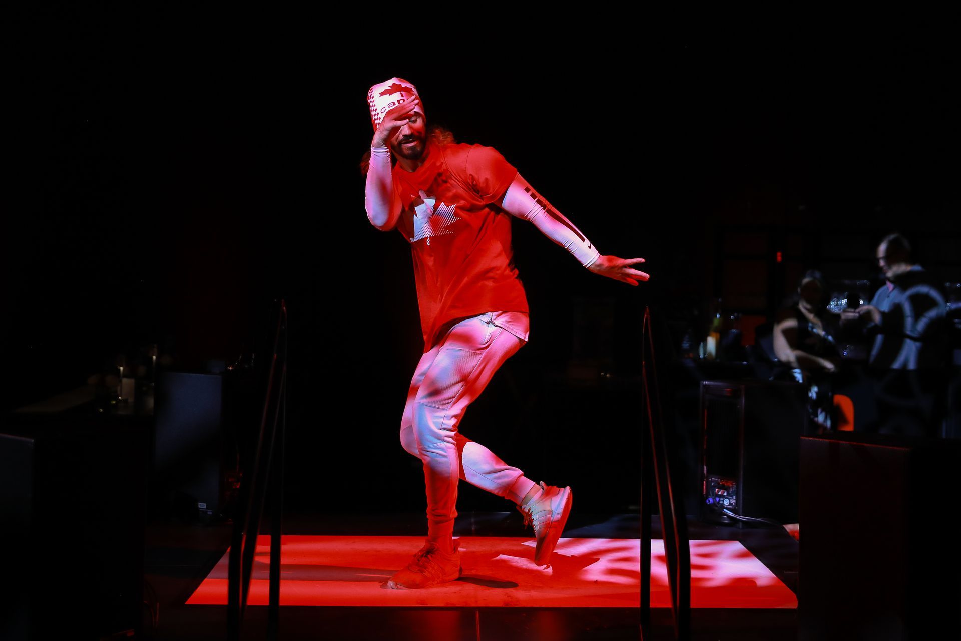 A person in red shirt and pants dances on a red lit stage, striking a pose with arm raised.