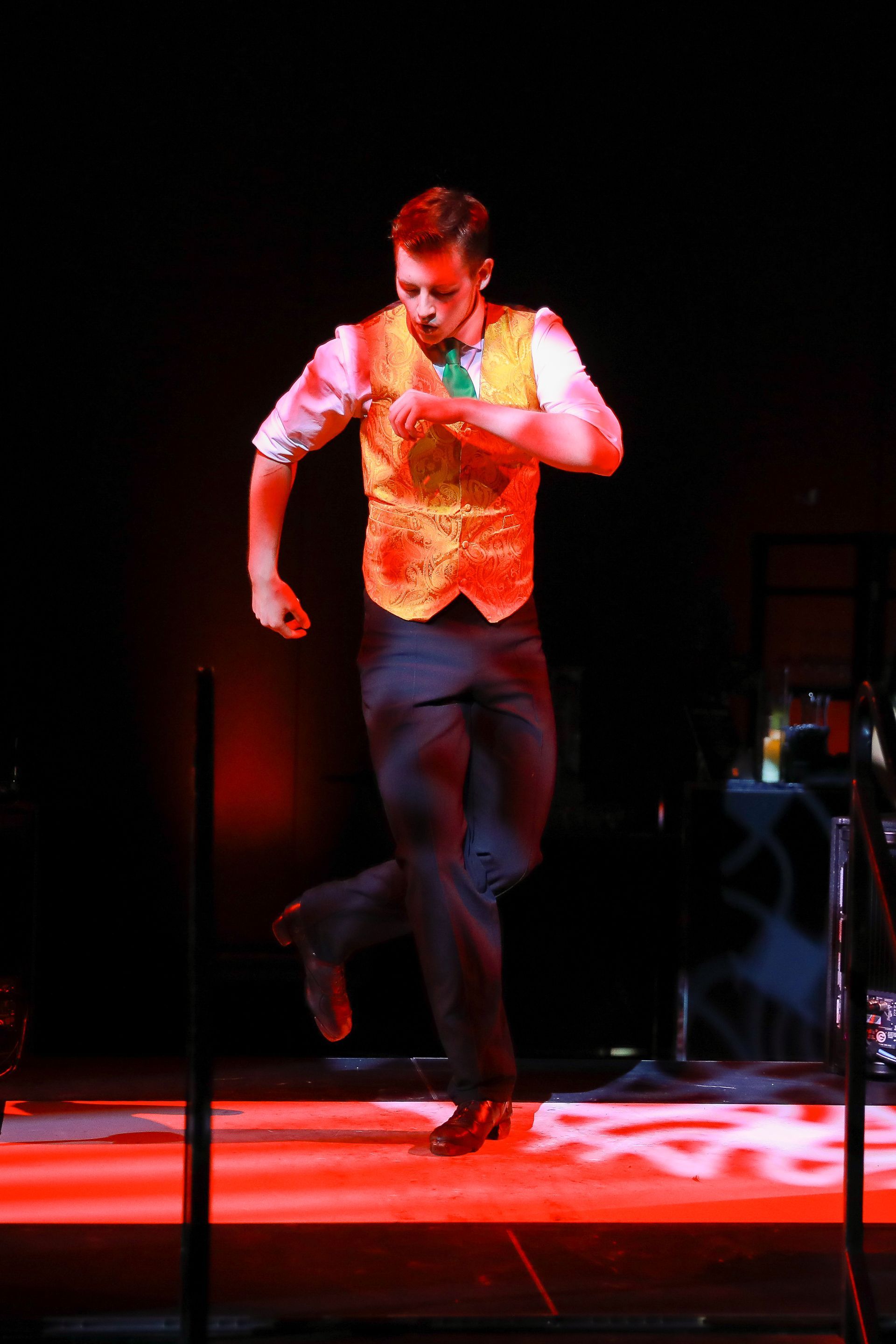 Man tap-dancing onstage, wearing a vest and tie, one leg raised, red lighting.