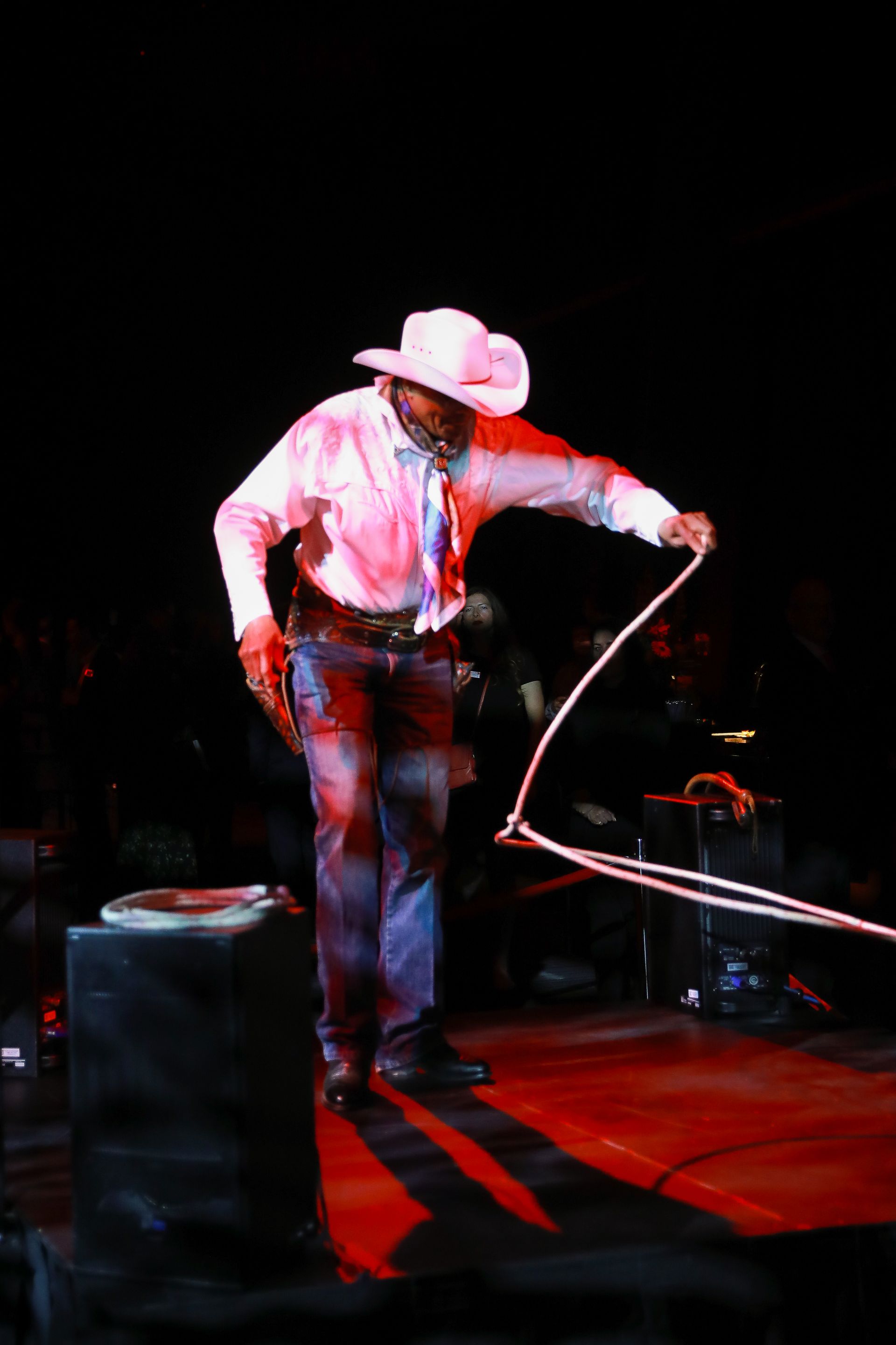 Cowboy in pink hat and shirt, twirling a lasso on a red-lit stage.