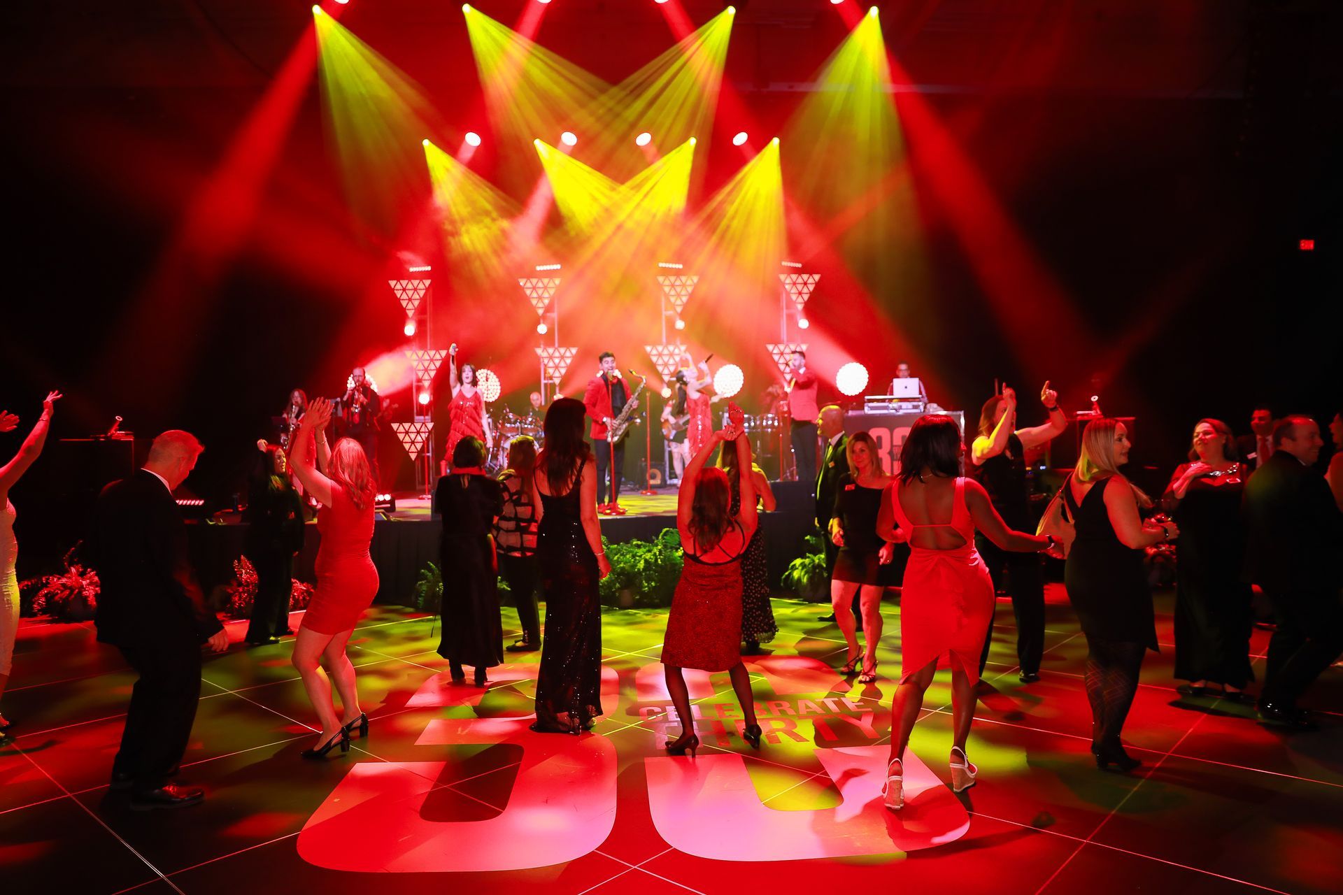 People dancing at a party with a band on stage, illuminated by red and yellow stage lights.